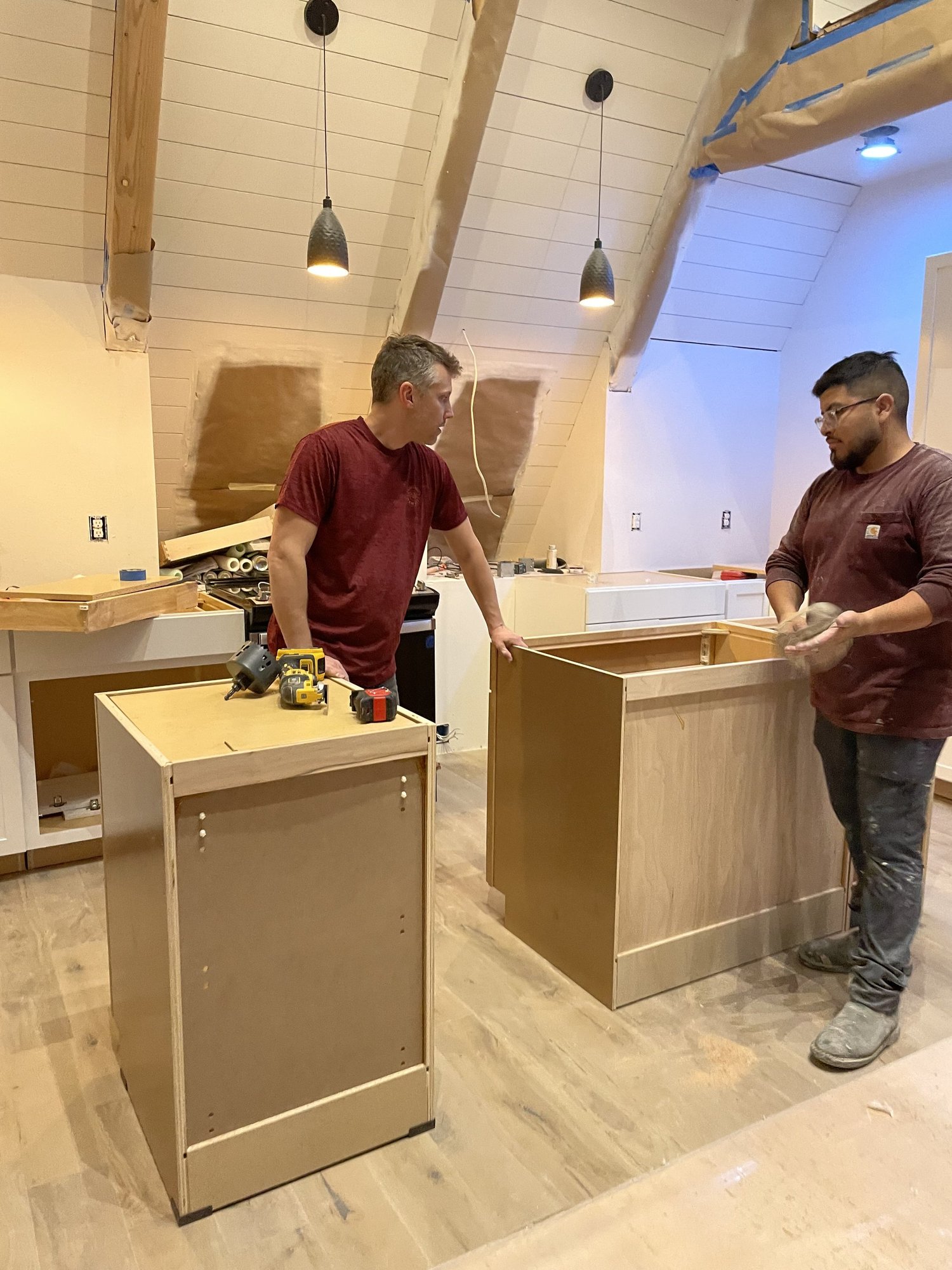 Two builders assembling kitchen cabinets under a wood-paneled ceiling